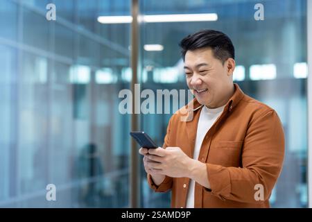Successful Asian businessman standing near the window with phone in hands. Man using application on smartphone, typing message and browsing social media inside office at workplace. Stock Photo