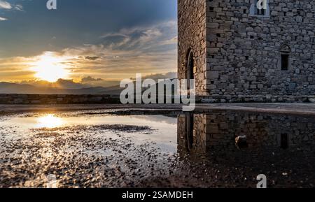 A scenic view of a stone tower at sunset, reflecting in a puddle on the ground. The sky is filled with clouds and warm colors, creating a tranquil atm Stock Photo