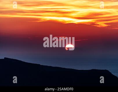 A stunning sunset over the ocean, with vibrant orange and yellow hues blending into the darkening sky. The sun is partially hidden behind clouds, cast Stock Photo