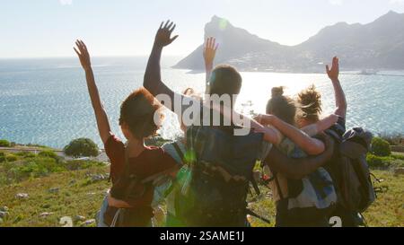 Rear View Of Group Of Young Friends Hiking Standing On Cliffs By Coast And Looking At Beautiful View Stock Photo