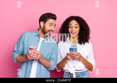 Excited young couple using smartphones against pink background, sharing joyful moments Stock Photo