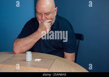 A man, seemingly distressed, sits at a table with a pill bottle and scattered pills.  The pensive posture and the presence of medication is a concern Stock Photo