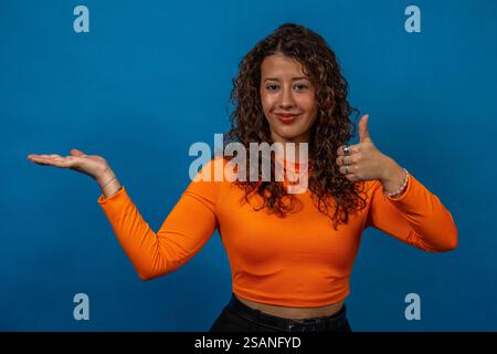 Studio portrait of a young woman gesturing thumbs up and holding her hand out, against a blue background Stock Photo