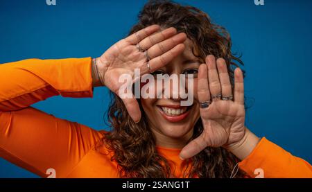 Cheerful young woman framing her face with hands, smiling and wearing an orange shirt on a vibrant blue background Stock Photo