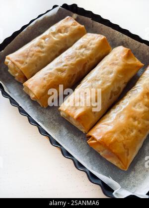 Freshly baked stuffed phyllo dough rolls in a baking pan on a white wooden table. Culinary, cooking, cooking concept. Directly above. Stock Photo