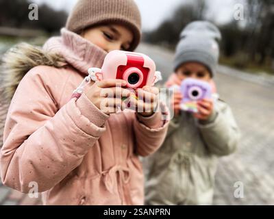 Two children enjoy the outdoors while playing with colorful toy cameras. They are dressed warmly, embracing creativity, play, and fun in a winter land Stock Photo