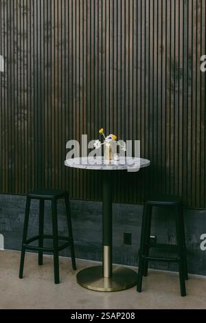 A small round table is adorned with a vase of fresh flowers, set against a stylish wooden wall. Two black stools flank the table, creating a welcoming Stock Photo