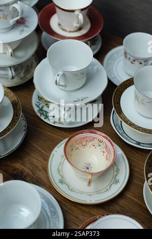A diverse assortment of vintage tea cups and saucers is displayed on a wooden surface. Each piece features unique designs and colors, highlighting int Stock Photo