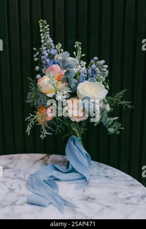 Delicate bouquet featuring roses, eucalyptus, and blue flowers sits elegantly on a marble table. A soft blue ribbon drapes alongside the arrangement, Stock Photo