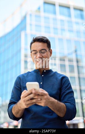 Happy Hispanic business man using mobile cell phone standing in city. Vertical Stock Photo