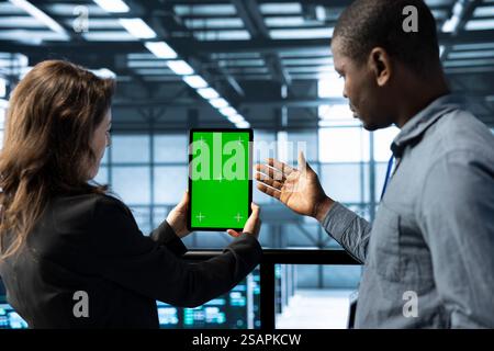 Team of engineers working in server farm, using chroma key tablet to analyze data. Employees working together in data center using isolated screen device, ensuring system integrity and security Stock Photo