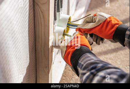 An electrician wearing safety gloves works on connecting electrical wires in a residential setting during daylight. The focus is on wiring processes. Stock Photo