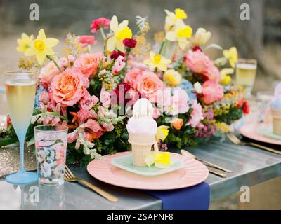 A vibrant outdoor table is beautifully arranged with a floral centerpiece featuring pink and yellow daisies, along with elegant glassware and a desser Stock Photo