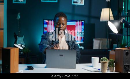 Woman arriving in apartment office, sitting down at desk, opening laptop to begin doing tasks. Teleworker starting job shift at home, preparing notebook device, logging in online, camera A Stock Photo