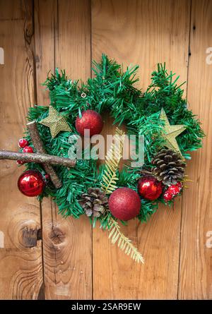 A festive Christmas wreath with green foliage, red baubles, pine cones, and gold stars, hanging on a wooden wall. Stock Photo