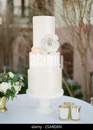 A three-tiered white wedding cake adorned with a large white flower stands on a table. Surrounding it are delicate candles and a small bouquet, enhanc Stock Photo
