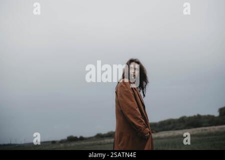 A woman in a brown coat stands distantly in a field under a calm, cloudy sky Stock Photo