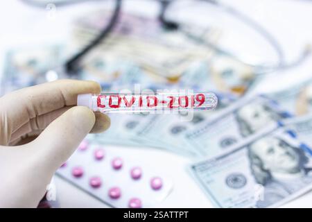 Gloved hand holding test tube labeled COVID-2019 amidst dollar bills, pills, and stethoscope Stock Photo