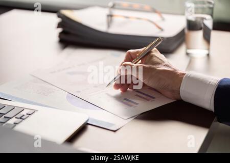 Elderly businessman analyzing financial reports and charts while taking notes at a modern office desk. Stock Photo