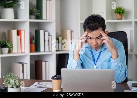 Stressed Asian businessman focusing on a laptop, dealing with challenges at his desk in a modern office environment. Stock Photo