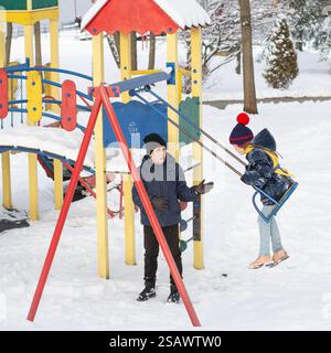 Two children enjoy a snowy winter day at an outdoor playground. One child pushes the other on a swing. The playground is brightly colored and partiall Stock Photo