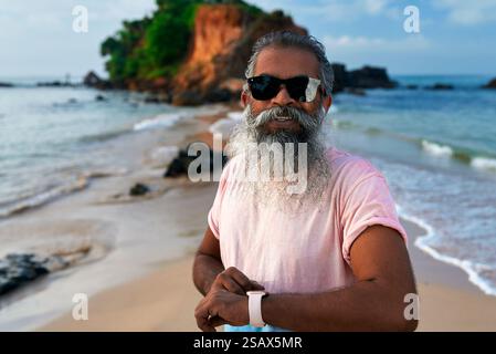 Senior South Asian man smiles on sunny beach. Wearing smart watch and earbuds. Bearded elderly using modern tech, fit lifestyle. Older adult embraces Stock Photo