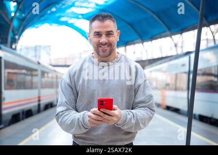 Smiling Man Using Red Smartphone at Train Station looking at camera Stock Photo