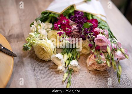 Beautiful assortment of vivid flowers wrapped in paper with a yellow-handled scissors nearby on a wooden surface, depicting creativity and preparation Stock Photo