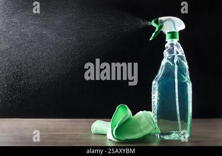 Transparent spray bottle with green liquid for cleaning expelling drops of product into the air on a wooden table with a green microfiber cloth and bl Stock Photo