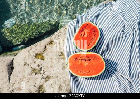 Freshly sliced watermelon on striped fabric beside crystal-clear water and rocky surface, creating vibrant summer vibes, natural textures, and refresh Stock Photo