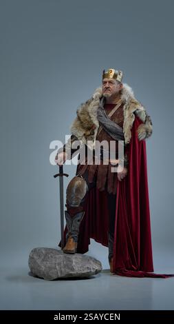 Senior serious man as medieval king standing on stone, leaning on sword with determination against grey studio background Stock Photo