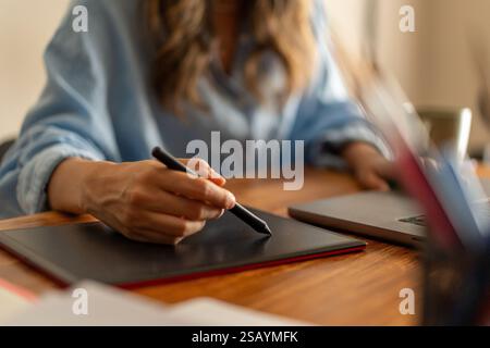 Close-up of a woman using a graphic tablet for digital design, working remotely from home with a laptop on a wooden desk. Stock Photo