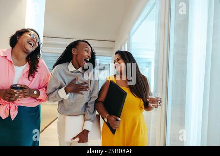 Three happy colleagues enjoying an excited conversation on a Friday afternoon, walking through a bright office hallway, sharing laughter and showcasin Stock Photo
