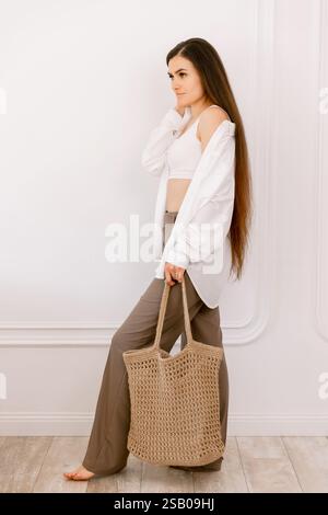 Elegant woman with long hair posing barefoot, carrying a stylish woven tote bag, showcasing casual chic fashion indoors Stock Photo