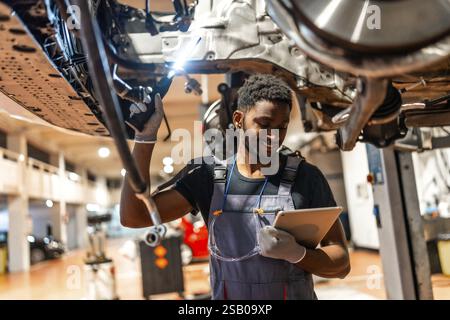 Young mechanic using a digital tablet while working on a car held up by a hydraulic lift in a professional auto repair shop Stock Photo