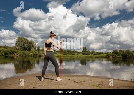 Woman working out on a riverbank using a resistance band, surrounded by blue sky and clouds Stock Photo