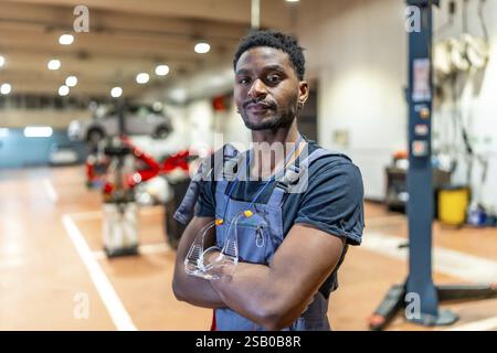 Young african american mechanic standing with crossed arms and protective eyewear in a car repair shop, exuding professionalism Stock Photo