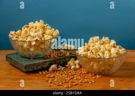 Two glass bowls filled with freshly popped popcorn surrounded by scattered corn kernels on a rustic wooden table, featuring a blue background. Stock Photo