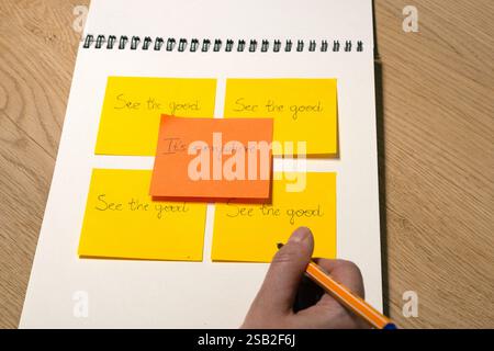 A person is writing on a yellow sticky note with a pen "See the good. It's everywhere." The paper has four yellow sticky notes on it.. Concept photo. Stock Photo