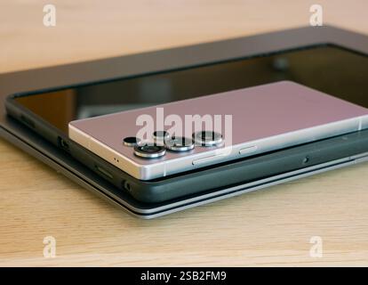 A new silver cell phone sits on top of a black tablet and tablet sits on silver laptop. The laptop is on a wooden table. Stock Photo