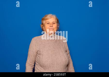Horizontal portrait of a Caucasian woman in her 70s posing proudly and satisfied with the stage of her life she is living, isolated on a blue studio Stock Photo
