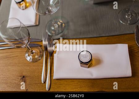 Elegant table setting with a small honey jar as a guest gift, placed on a folded white napkin, with silverware and wine glasses on a wooden table Stock Photo