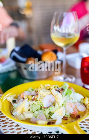 A Caesar salad served on a yellow plate, topped with shrimp, croutons, and egg, accompanied by a glass of white wine at a restaurant table Stock Photo