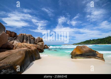 Scenic view of a beach in Seychelles, featuring large granite rock formations, clear turquoise water, white sand, and a blue sky with scattered clouds Stock Photo