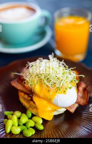 A gourmet serving of Eggs Benedict topped with fresh sprouts and creamy hollandaise sauce, accompanied by edamame, with a cappuccino and orange juice Stock Photo