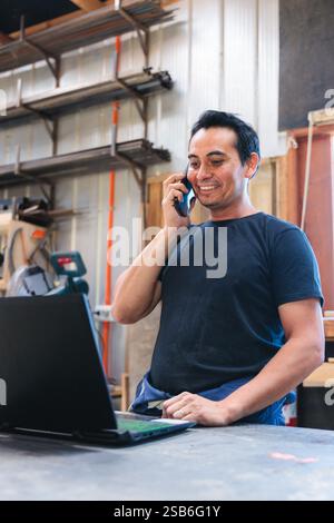 Craftsman managing orders using laptop and talking on phone in workshop Stock Photo