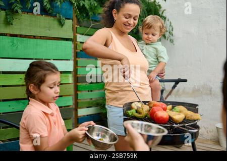 A cheerful mother cooks vegetables on a grill while children assist during an outdoor family barbecue. Stock Photo