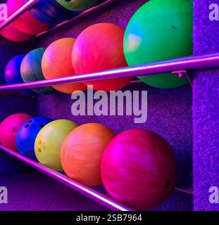 Bowling balls of various colors are neatly arranged on a metal rack, illuminated by vibrant purple light, ready for bowlers to select and enjoy a game Stock Photo