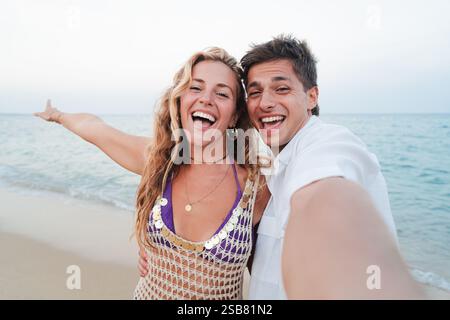 Laughing couple taking a selfie on the beach, woman pointing joyfully at the ocean while man holds camera, capturing their happiness and playful moment under the bright sun. High quality photo Stock Photo