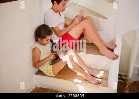 Two children sitting on stairs using digital devices, focusing on tablets and smartphones. Stock Photo